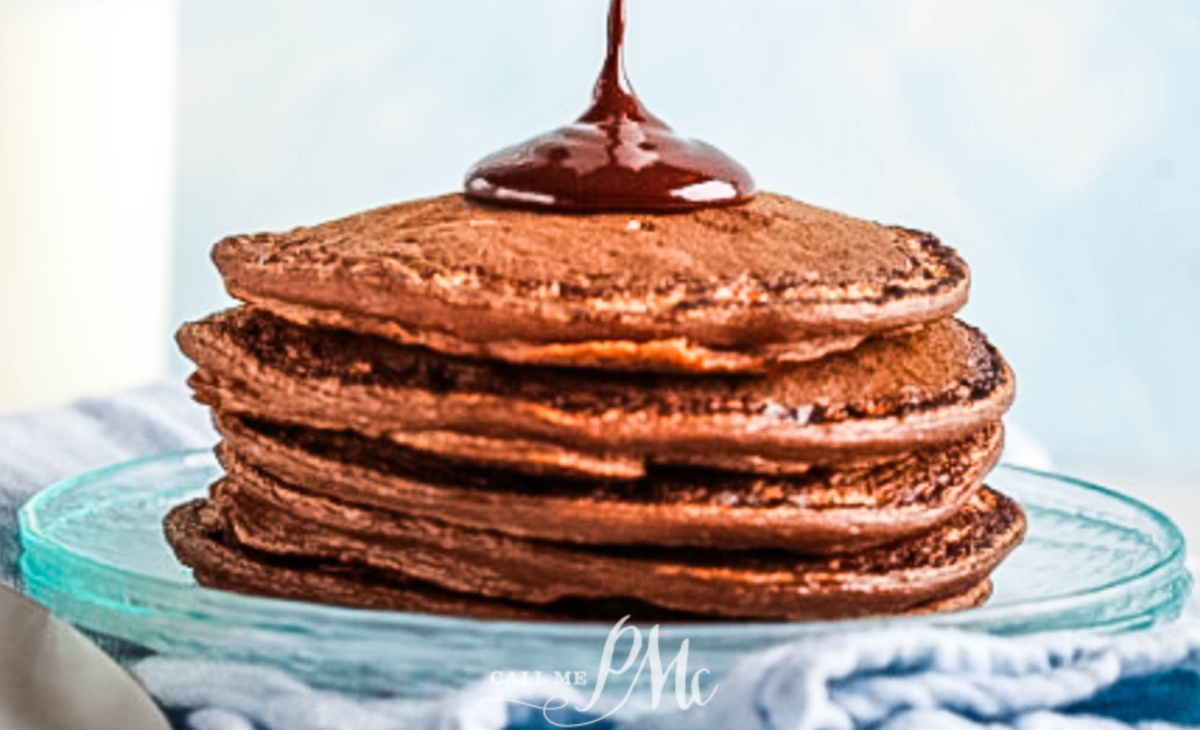A stack of Chocolate Chocolate Chip Pancakes on a glass plate, topped with rich chocolate syrup being poured over them, with a light blue background and a blue striped cloth nearby.