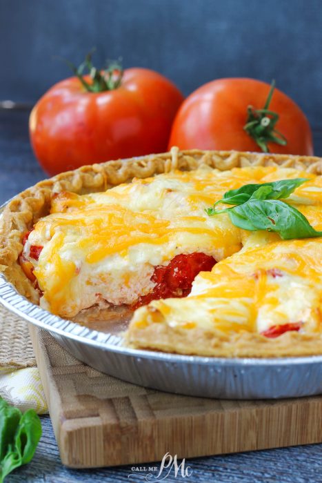 A Southern Tomato Pie in a metal pan, topped with melted cheese and basil leaves, features sliced tomatoes and cheese, while whole tomatoes sit in the background.