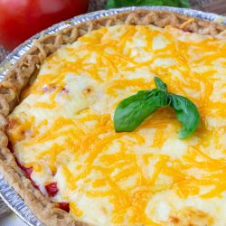 A classic Southern Tomato Pie topped with melted cheese and garnished with fresh basil, served in a foil pie dish, with ripe tomatoes and basil in the background.
