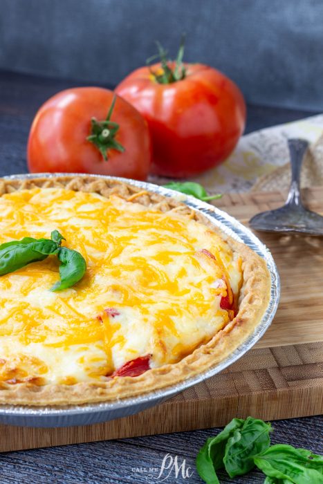 A baked cheesy Southern Tomato Pie in a foil dish is garnished with basil, with fresh tomatoes and basil leaves on a wooden cutting board in the background.