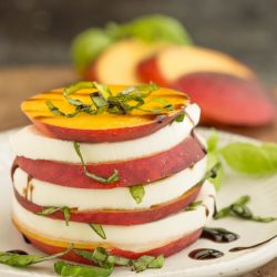 Enjoy a refreshing Peach Caprese Salad with Balsamic Reduction, featuring stacked peach and mozzarella slices topped with basil, all drizzled elegantly on a white plate. In the background, extra peach slices and basil leaves enhance the presentation.