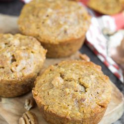 Three homemade pecan pie muffins sit on a wooden board, surrounded by scattered pecan pieces. The muffins have a golden-brown, slightly textured top.