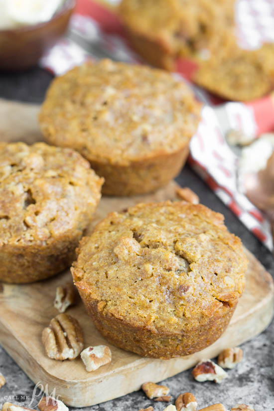 Three homemade pecan pie muffins sit on a wooden board, surrounded by scattered pecan pieces. The muffins have a golden-brown, slightly textured top.