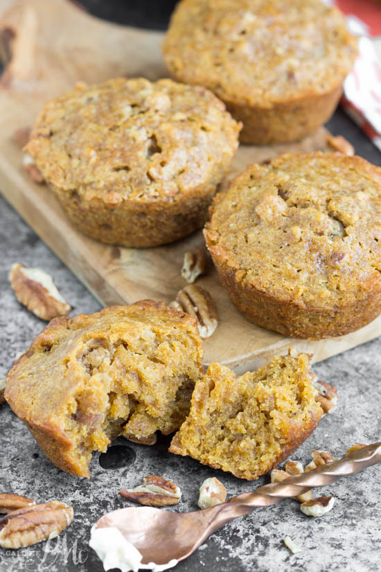 Four pecan pie muffins, one broken in half, sit on a wooden board with scattered pecans around them on a gray surface.