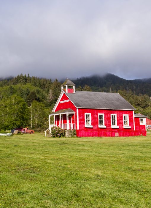 Mom's Survival Guide to Starting School A bright red schoolhouse with a bell tower sits on a grassy field, surrounded by trees and hills under a cloudy sky—a classic scene right out of Mom's Survival Guide to Starting School.