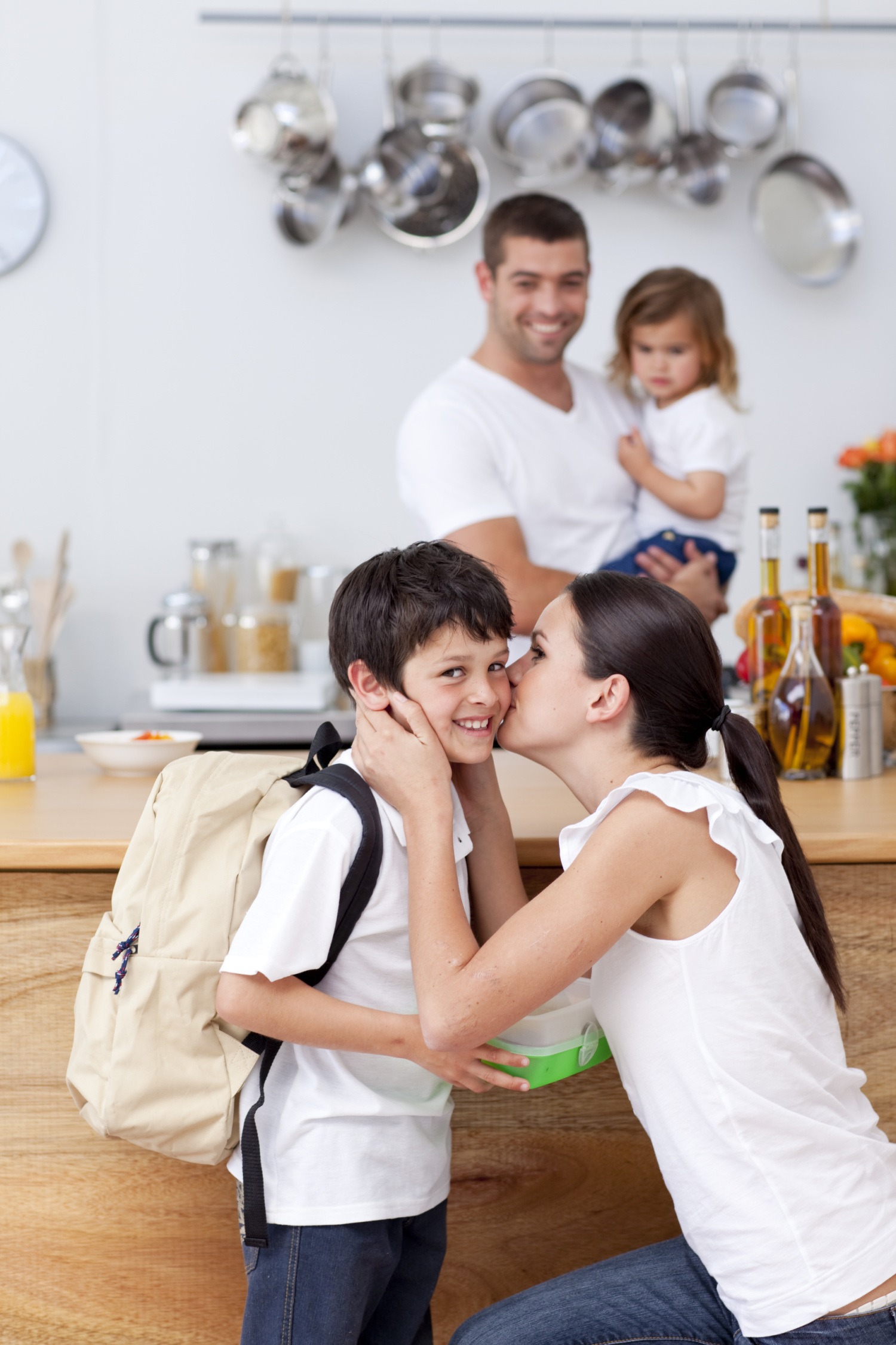 A woman kisses a boy wearing a backpack, while a man holds a young child in the kitchen—capturing the spirit of Mom's Back to School Survival Guide.