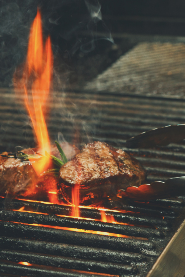 Two steaks on the grill with flame and tongs.