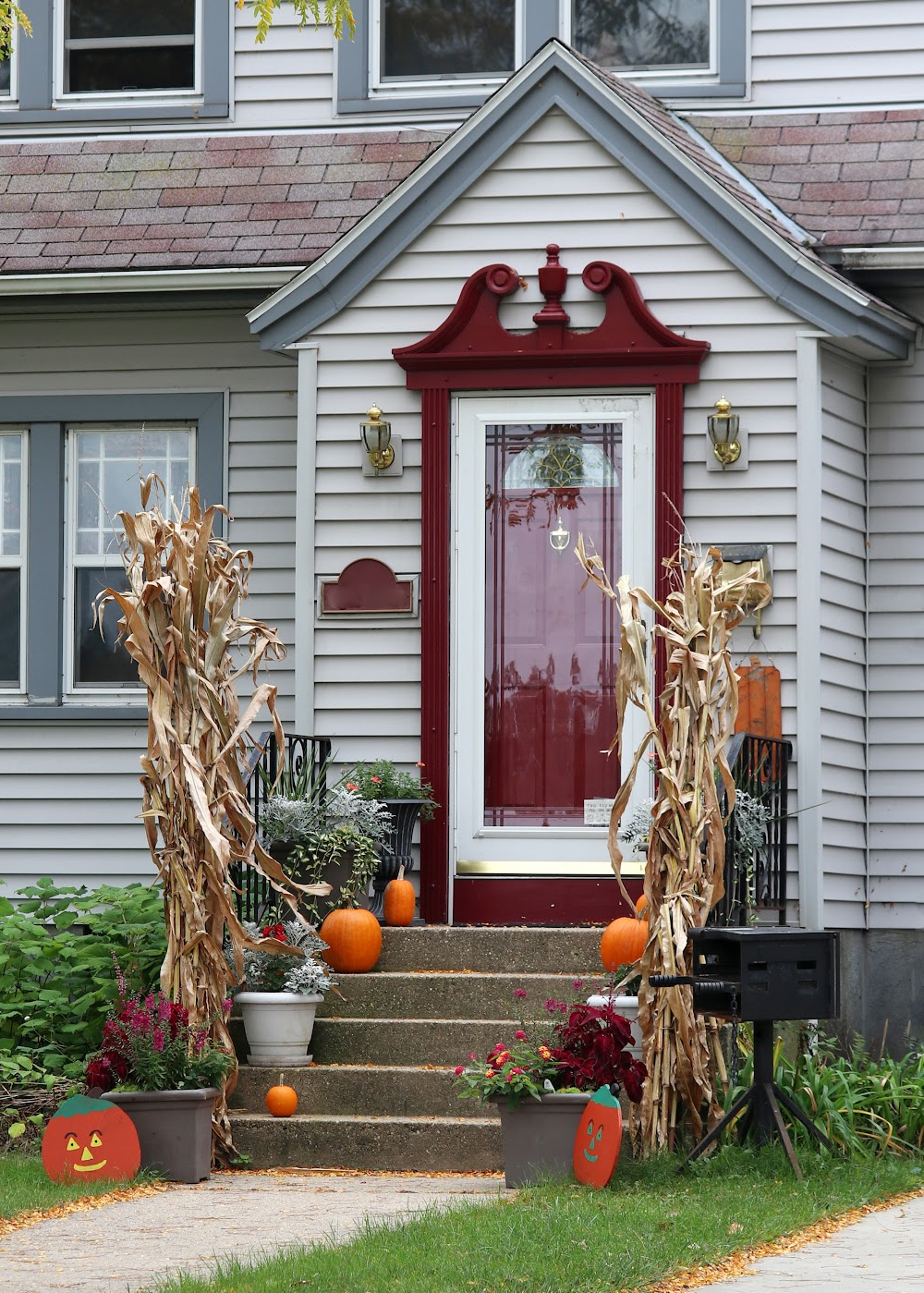Front porch decorated for autumn, adding fall color with cornstalks, pumpkins, potted plants, and Halloween-themed signs by the steps of a gray house with a red door.