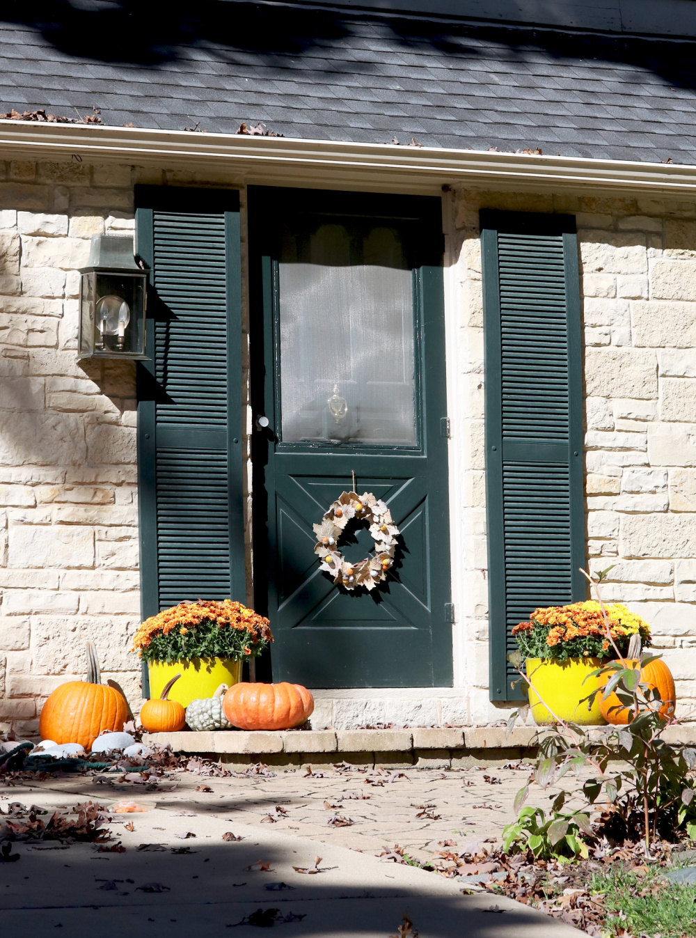 A dark green door with a wreath, flanked by green shutters, two planters with orange flowers, and several pumpkins on a stone porch—adding fall color to the entryway.