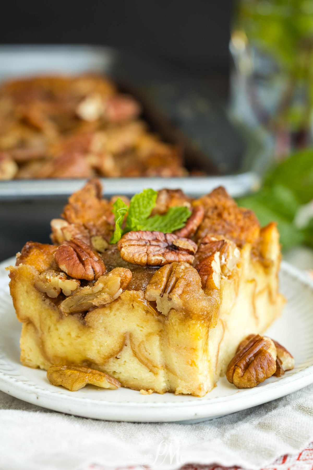 A close-up of Pecan Pie Bread Pudding topped with pecans and a sprig of mint, served on a white plate. More bread pudding is blurred in the background.