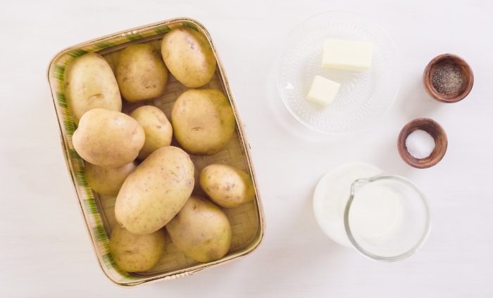 A basket of Parmesan Potatoes, a glass pitcher of milk, a plate with two pats of butter, and small bowls of salt and pepper are arranged on a white surface.
