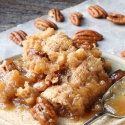 A plate of pecan pie bread pudding with caramel sauce and scattered pecans evokes the rustic charm of a pecan cobbler, all beautifully arranged on a wooden surface.