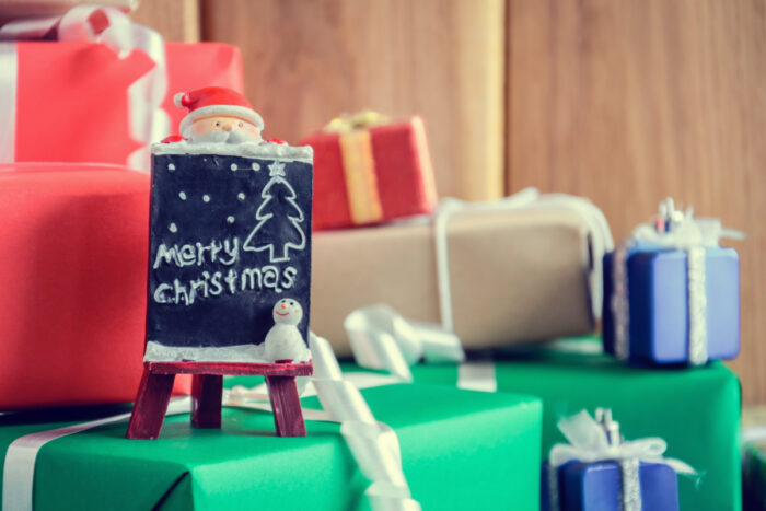 A small chalkboard with a Santa figurine and “Merry Christmas” written on it is surrounded by wrapped gifts in red, green, brown, and blue paper with white ribbons, set against a wooden background—creating a festive scene reminiscent of The Happy Elf’s holiday workshop.