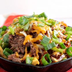 A skillet brimming with Irish Pub-Style Potato Nachos, featuring shredded beef, melted cheese, and chopped green onions, all served on a red and white checkered cloth.