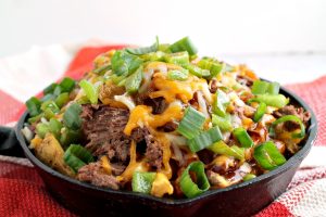 A skillet brimming with Irish Pub-Style Potato Nachos, featuring shredded beef, melted cheese, and chopped green onions, all served on a red and white checkered cloth.