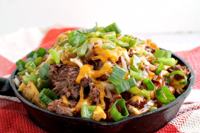 A skillet brimming with Irish Pub-Style Potato Nachos, featuring shredded beef, melted cheese, and chopped green onions, all served on a red and white checkered cloth.