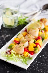 Tangy Teriyaki Scallops seared to perfection, served on a bed of mixed diced vegetables and mango, garnished with lime slices and fresh greens, with a glass of water and utensils in the background.