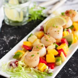 Tangy Teriyaki Scallops seared to perfection, served on a bed of mixed diced vegetables and mango, garnished with lime slices and fresh greens, with a glass of water and utensils in the background.