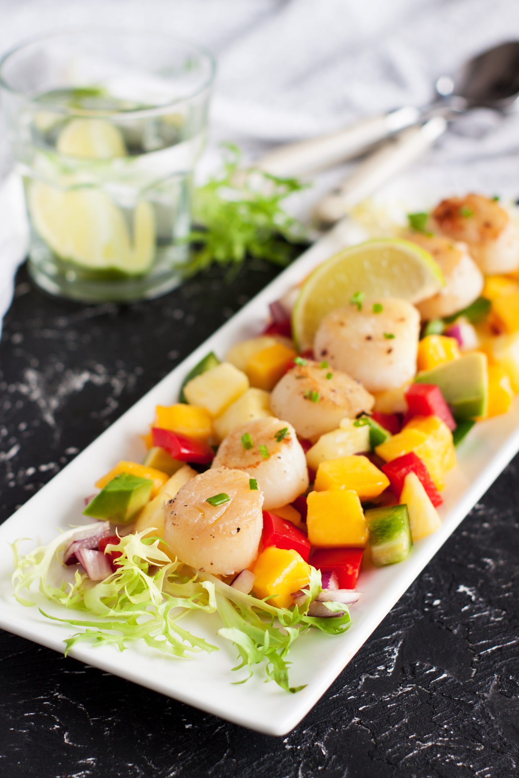 Tangy Teriyaki Scallops seared to perfection, served on a bed of mixed diced vegetables and mango, garnished with lime slices and fresh greens, with a glass of water and utensils in the background.