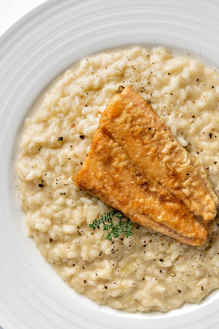 Pan Seared Catfish over Champagne Risotto is served on a white plate, garnished with a small sprig of fresh herb and black pepper for an elegant finish.