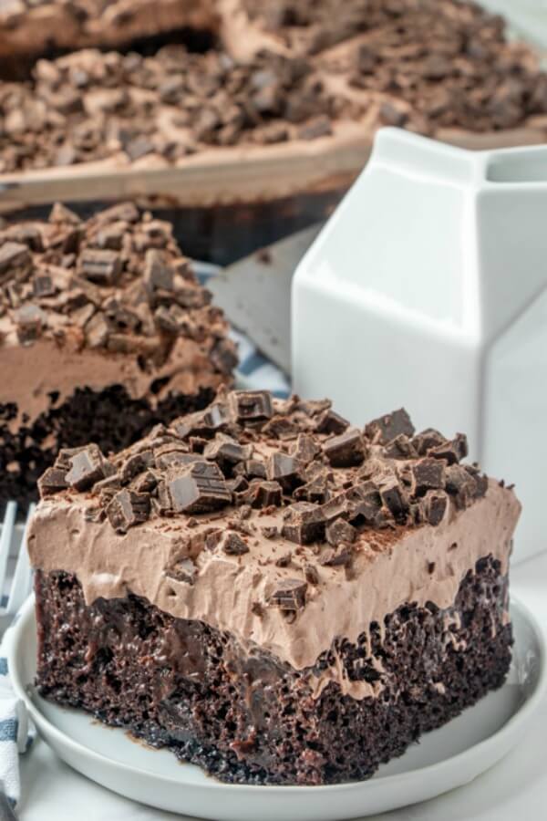 Death By Chocolate Poke Cake A slice of 50 Shades of Chocolate cake with chocolate frosting and chocolate chunks on top sits on a plate, with a cake dish and a milk carton-shaped container in the background.