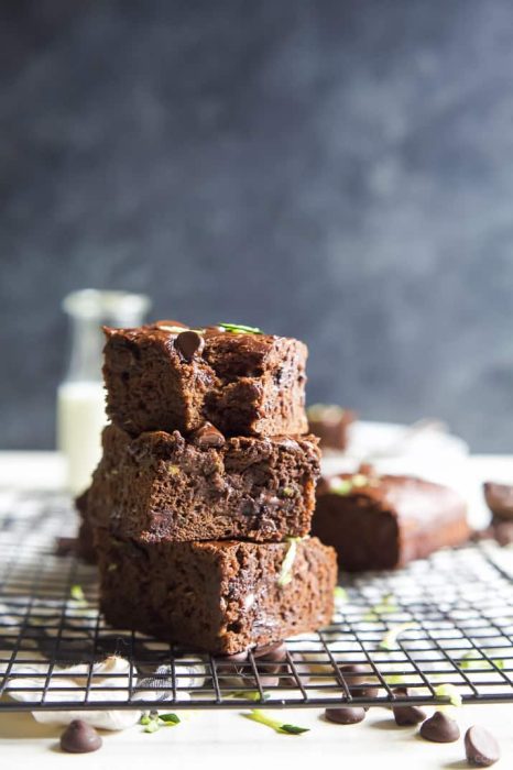Three 50 Shades of Chocolate brownies are stacked on a black cooling rack, with chocolate chips scattered around and a blurred glass of milk in the background.