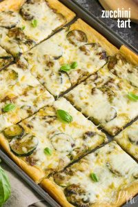 A fresh zucchini tart, baked to perfection and cut into rectangular pieces, is topped with melted cheese and garnished with aromatic basil leaves.