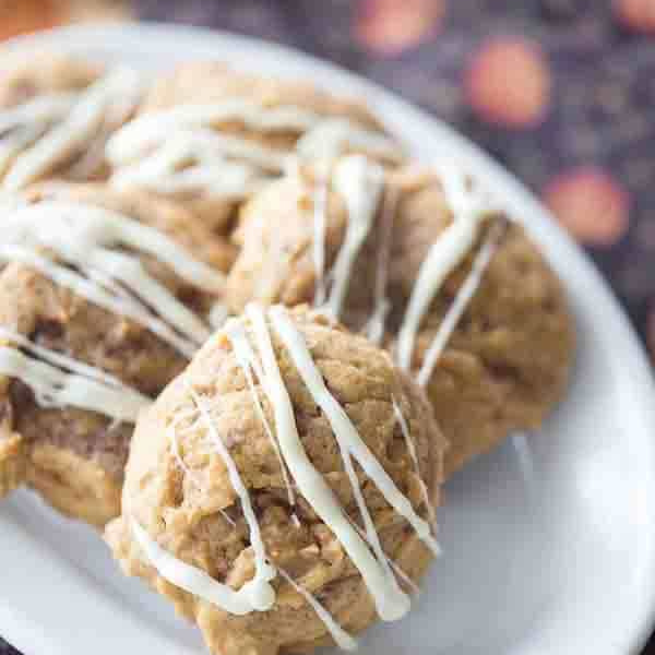 Three large cookies drizzled with white icing are arranged on a white plate, ready to bring a taste of The Great Pumpkin Palooza to your table. The cookies are golden brown with a soft texture, perfectly capturing the cozy spirit of the season. In the background, hints of a dark surface with orange spots add a festive touch to the blurred setting.