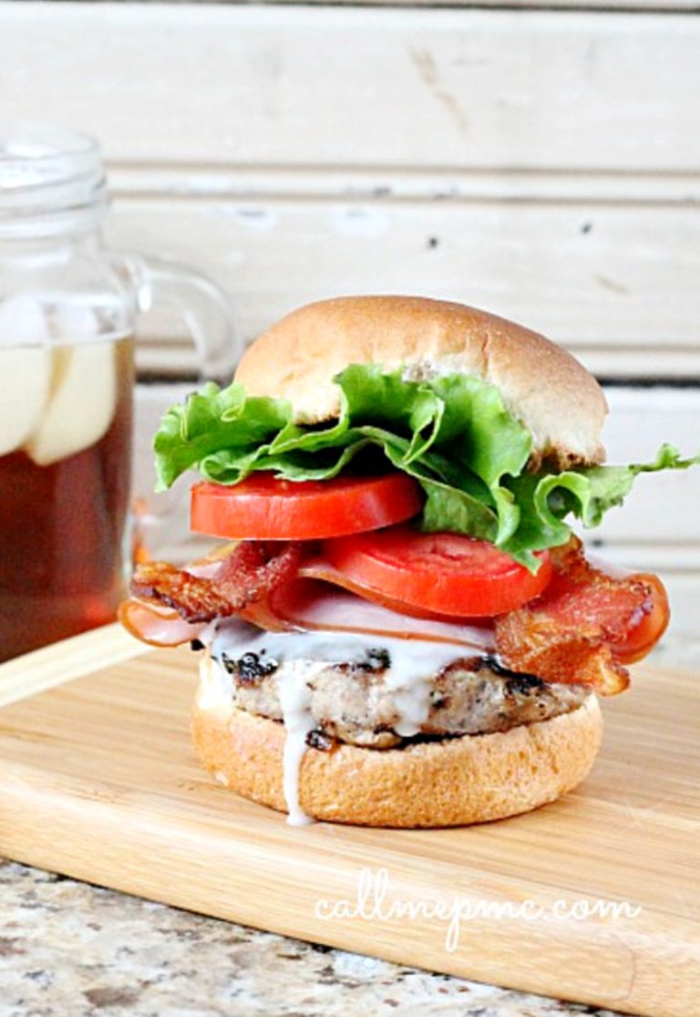 A Kentucky Hot Brown Burger with bacon, lettuce, tomato, and savory sauce sits enticingly on a wooden board. Nearby, a jar of refreshing iced tea completes this delightful scene.