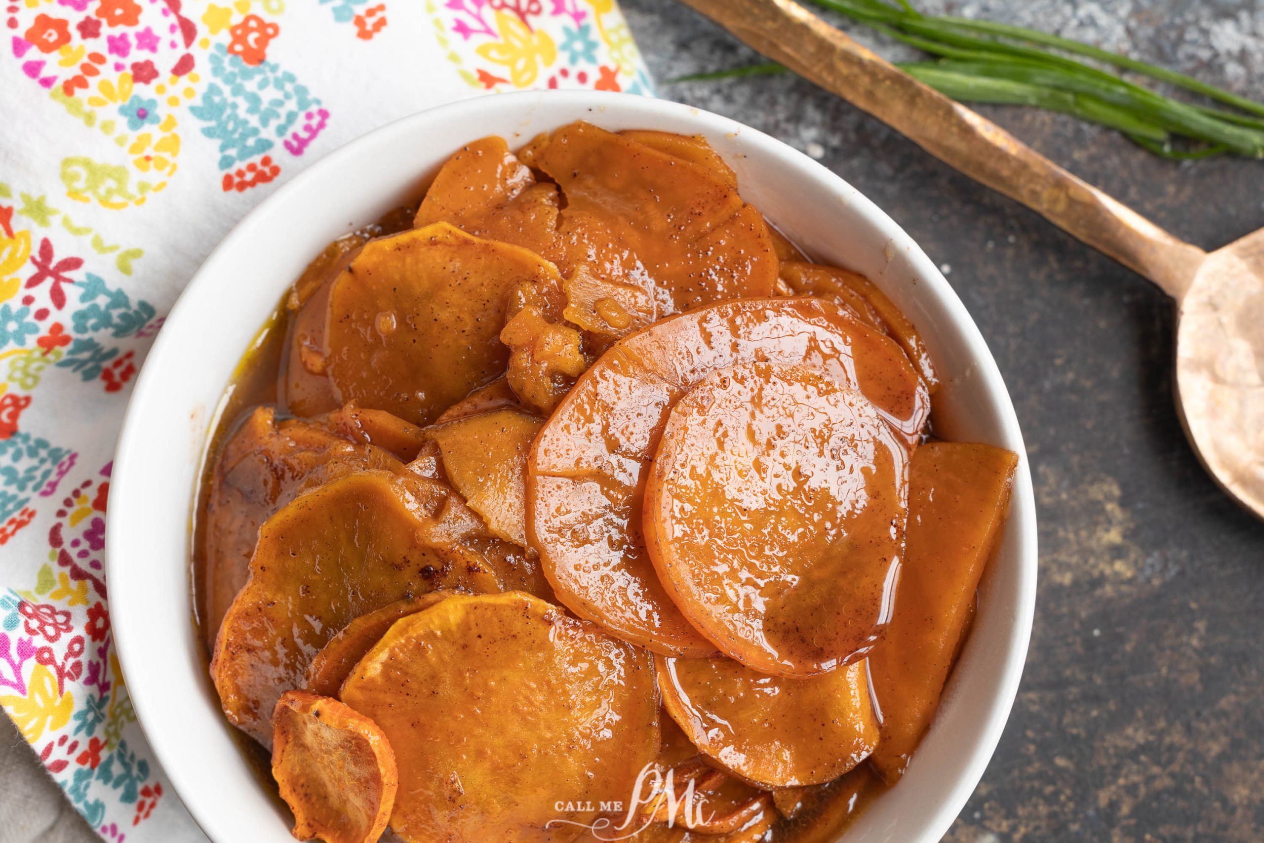 A white bowl filled with Thanksgiving Candied Sweet Potatoes, sliced and cooked in a brown, glossy sauce, sits on a dark surface next to a floral napkin and a copper spoon.