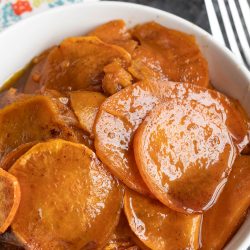 A white bowl filled with Thanksgiving Candied Sweet Potatoes—sliced, glazed in a rich brown syrup—with a fork and a colorful napkin nearby.