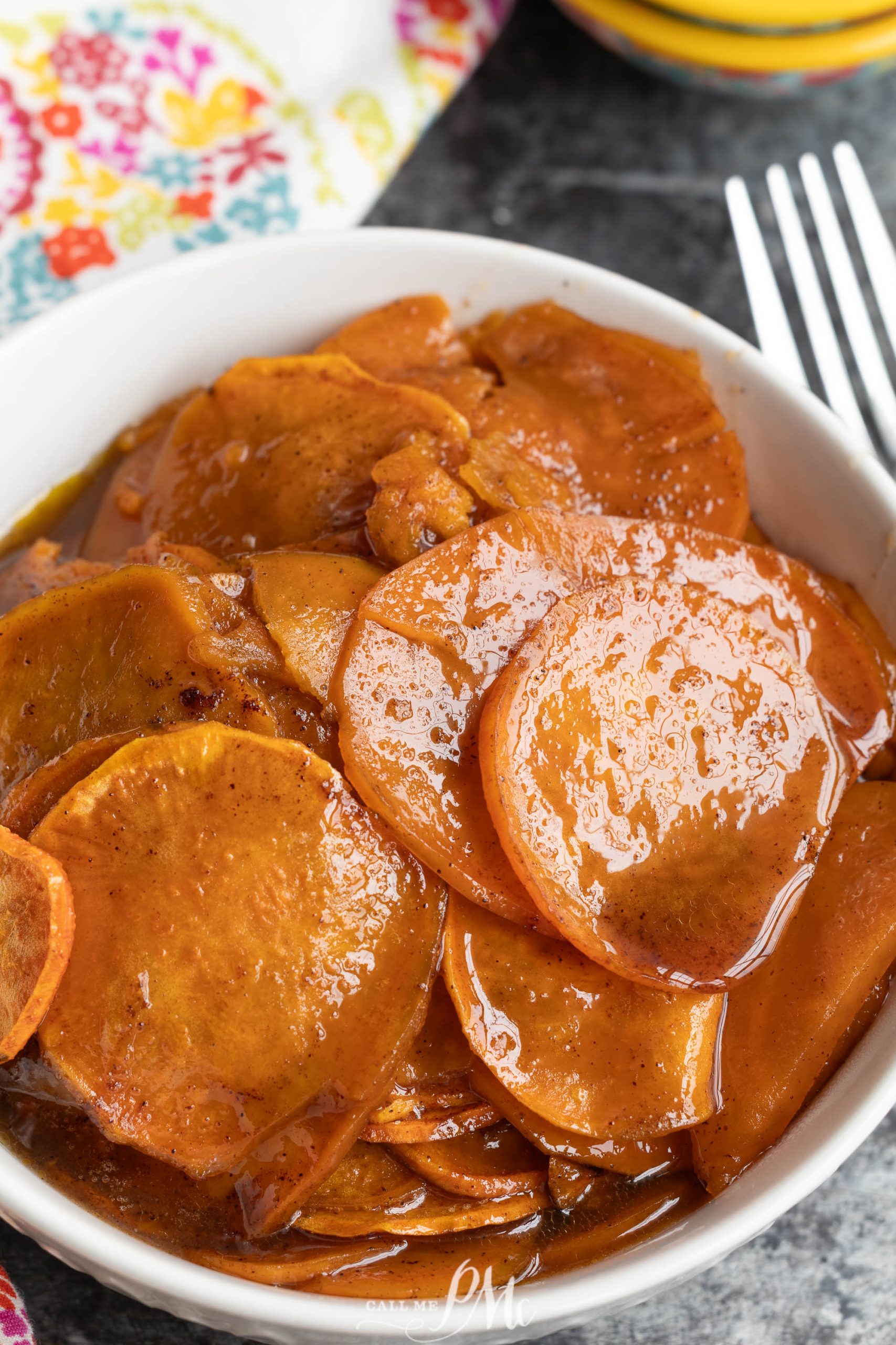 A white bowl filled with Thanksgiving Candied Sweet Potatoes—sliced, glazed in a rich brown syrup—with a fork and a colorful napkin nearby.