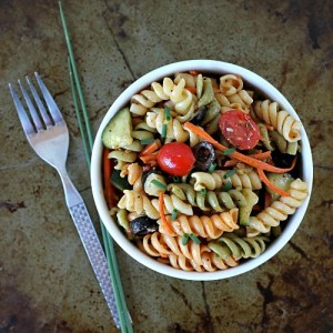 A bowl of vibrant vegetable pasta salad, featuring rotini intertwined with cherry tomatoes, cucumbers, carrots, and chives, sits invitingly next to a fork on a textured surface.