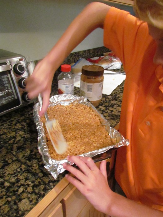 Young boy spreading peanut butter cereal bars mixture into a foil lined pan.