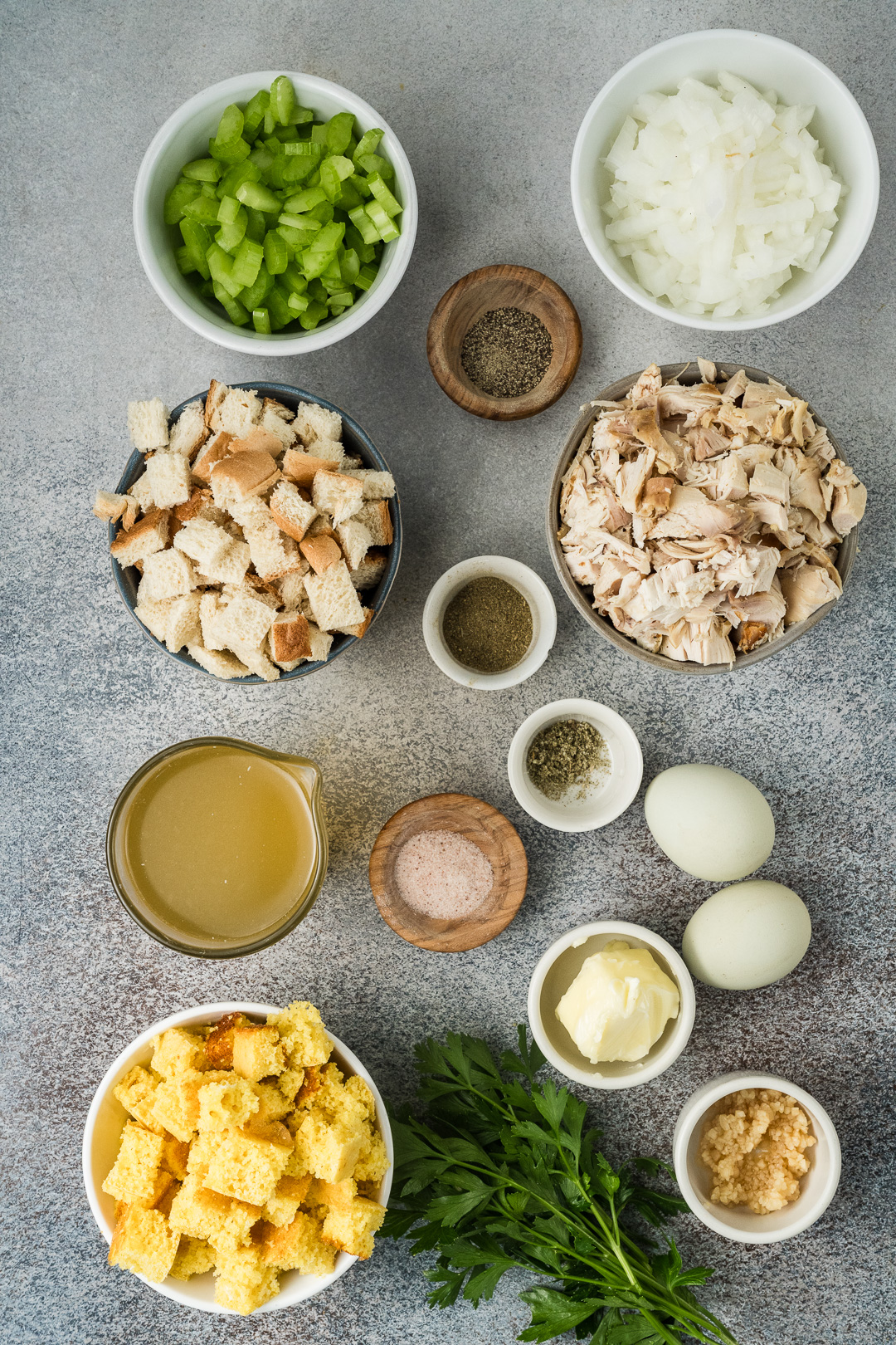 Ingredients for Southern cornbread dressing are arranged on a gray surface, including chopped celery, onion, bread cubes, shredded chicken, cornbread cubes, eggs, broth, parsley, butter, garlic, and various spices in small bowls.
