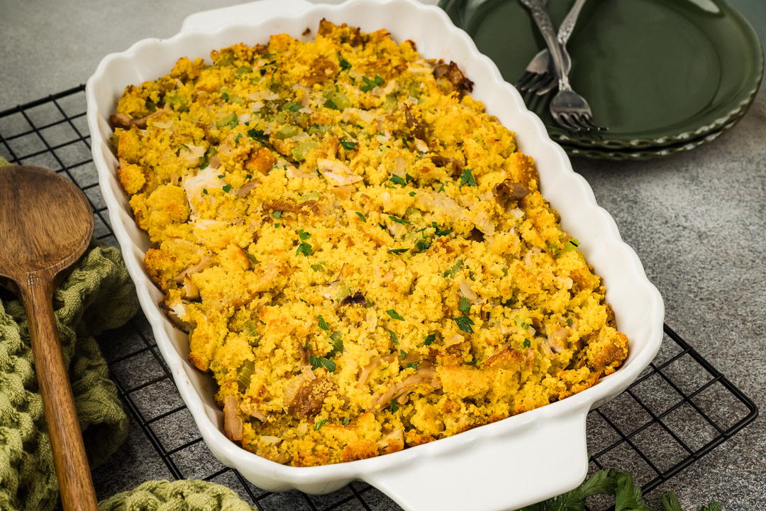 A white baking dish filled with golden brown Southern cornbread dressing sits on a cooling rack, with a wooden spoon, green napkin, and stacked green plates with forks nearby.