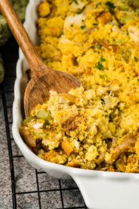 A wooden spoon serves Southern cornbread dressing, with visible celery and bread pieces, from a white baking dish on a cooling rack.