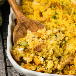 A wooden spoon serves Southern cornbread dressing, with visible celery and bread pieces, from a white baking dish on a cooling rack.