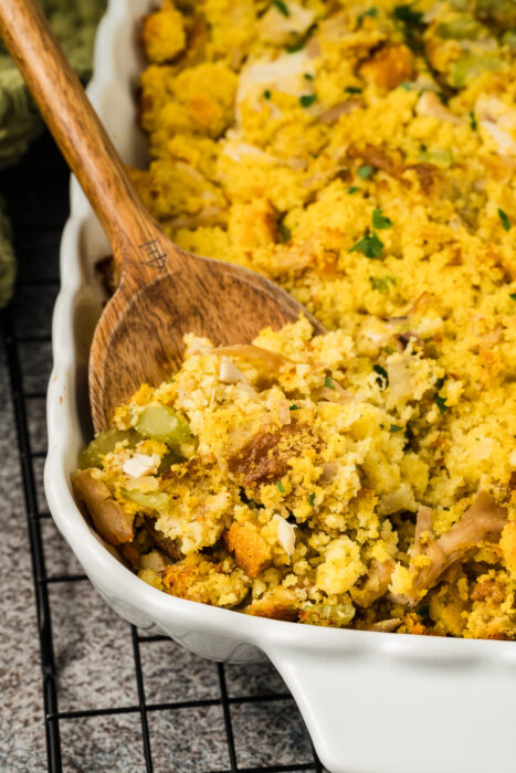 A wooden spoon serves Southern cornbread dressing, with visible celery and bread pieces, from a white baking dish on a cooling rack.