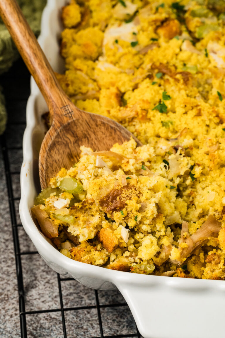 A wooden spoon serves Southern cornbread dressing, with visible celery and bread pieces, from a white baking dish on a cooling rack.