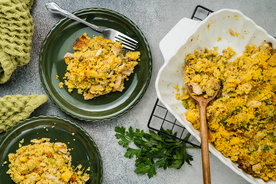 A serving of Southern cornbread dressing with chicken on a green plate, next to a baking dish with more dressing, a wooden spoon, parsley, and green knitted potholders on a gray surface.