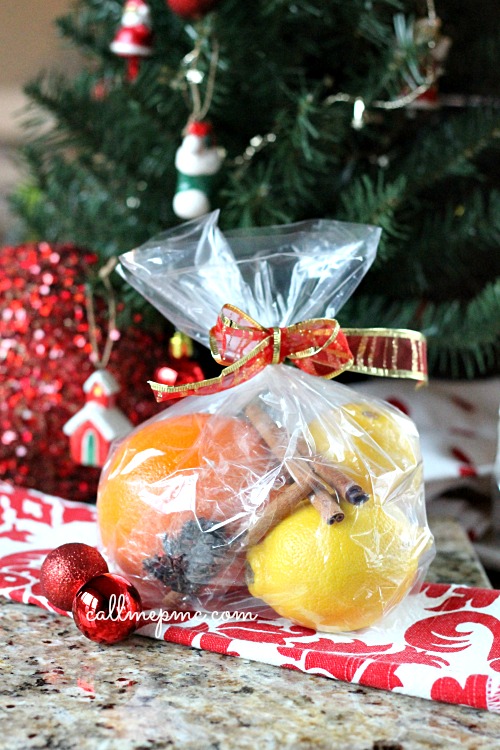 Simmering Holiday Potpourri A clear gift bag with simmering holiday potpourri, featuring oranges, a lemon, cinnamon sticks, and a pinecone tied with a ribbon. A Christmas tree twinkles in the background.