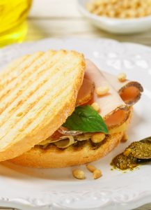 A toasted Baked Focaccia Turkey Sandwich with slices of turkey, basil, pine nuts, and cheese, served on a white plate with a spoonful of pesto on the side.