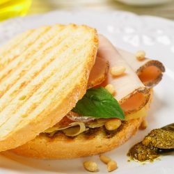 A toasted Baked Focaccia Turkey Sandwich with slices of turkey, basil, pine nuts, and cheese, served on a white plate with a spoonful of pesto on the side.