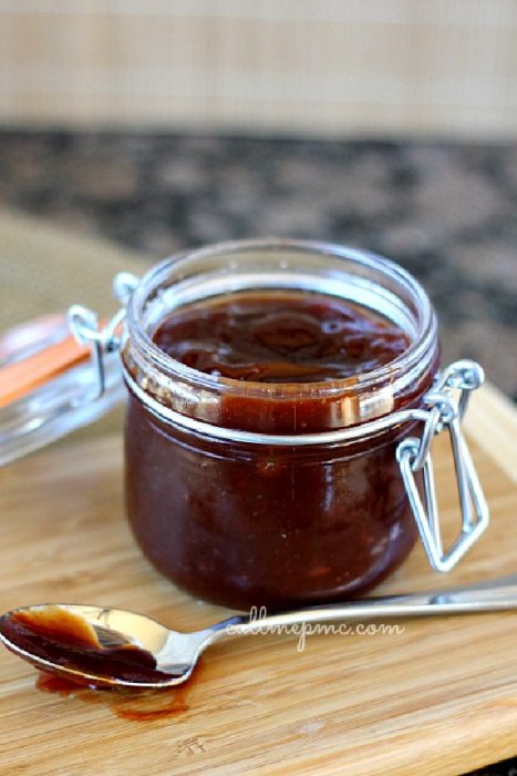 An open glass jar filled with dark brown Slow Cooker Barbecue Sauce sits on a wooden board, with a spoon covered in the same sauce placed beside it.