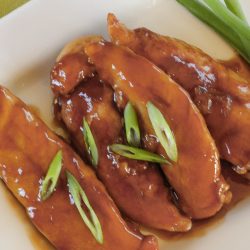 Three glazed chicken tenders, crafted from our Sticky Chicken Tenders Recipe and garnished with sliced green onions, are arranged on a white plate.