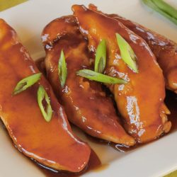 Three glazed chicken tenders, prepared using our Sticky Chicken Tenders Recipe, are topped with sliced green onions and served on a white rectangular plate.