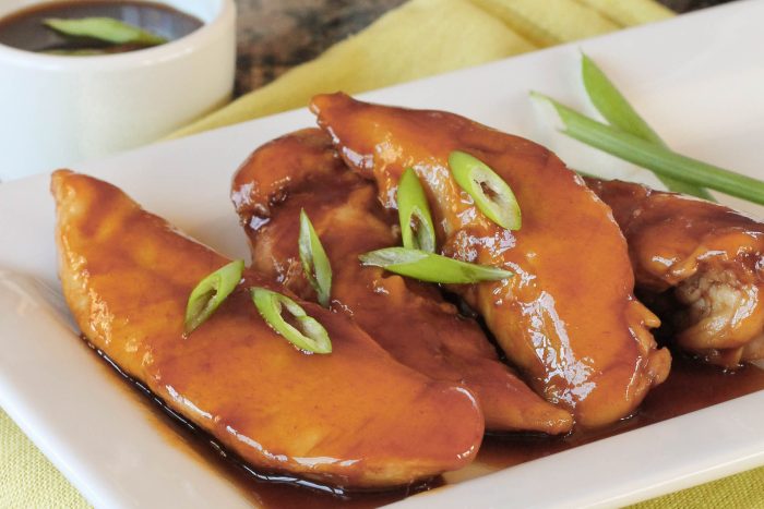 Three glazed chicken tenders from our Sticky Chicken Tenders Recipe, garnished with sliced green onions on a white plate, with a cup of sauce and green onion stalks in the background.