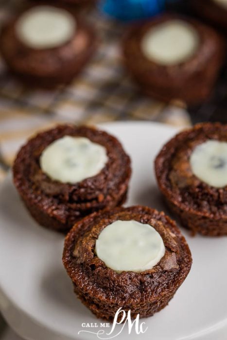 Brownie Kisses are rich, decadent chocolate fudge bites made more special crowned with cookies and cream chocolate Kiss while they're still hot and gooey. #brownies #recipes #cupcakes #cake #chocolate Three Brownie Kisses chocolate brownie cups with a white chocolate center sit on a white plate, surrounded by more brownies softly blurred in the background.