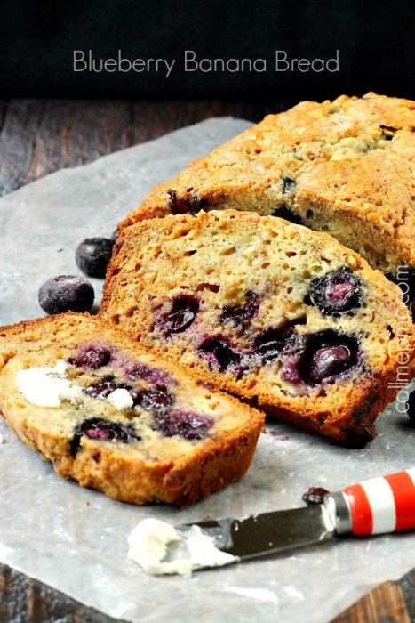 A sliced loaf of Blueberry Banana Bread with Almond Milk sits on parchment paper, with a buttered slice in the foreground and a red-handled knife nearby.
